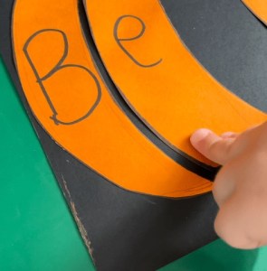 a partial orange pumplkin with the leters B and e. A child's hand is sticking the parts down