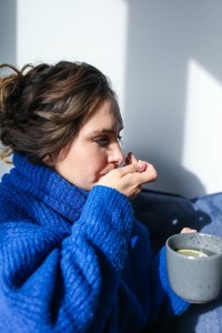 A woman wears a thick blue jumper and has a hot drink in one hand. The other hand is at her mouth as though she is taking medication.